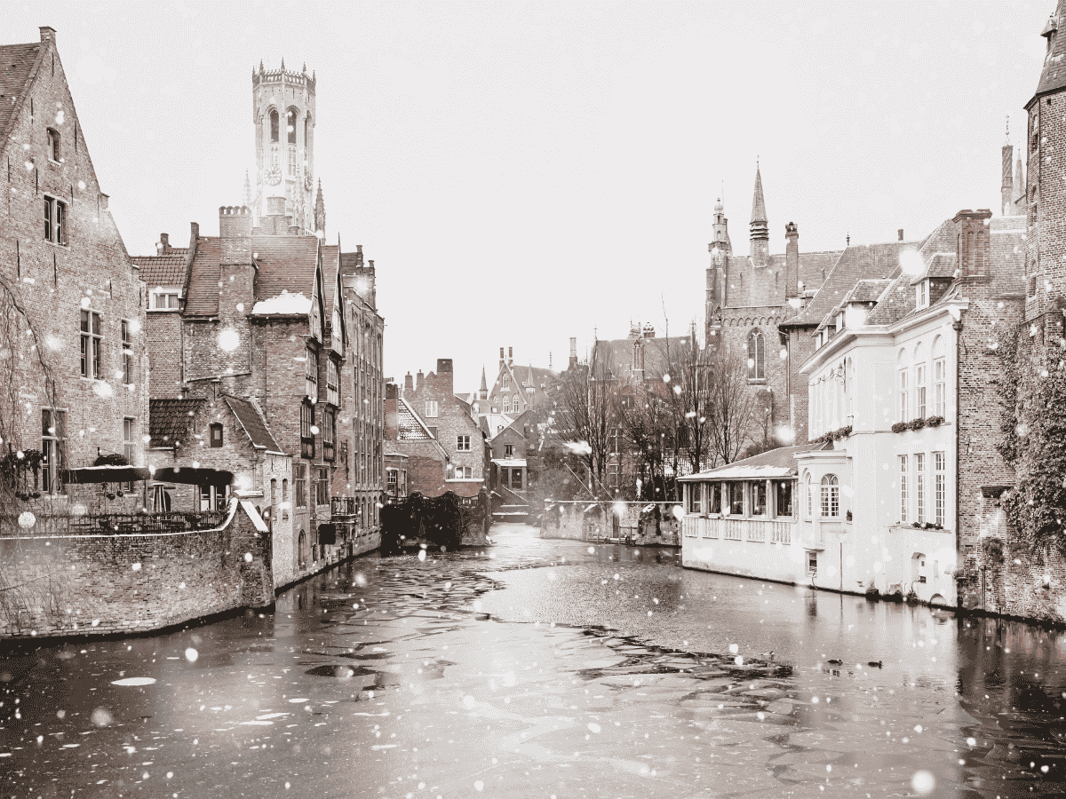 Snow falls softly over the frozen canals and historic brick buildings of Bruges, creating a serene winter scene.