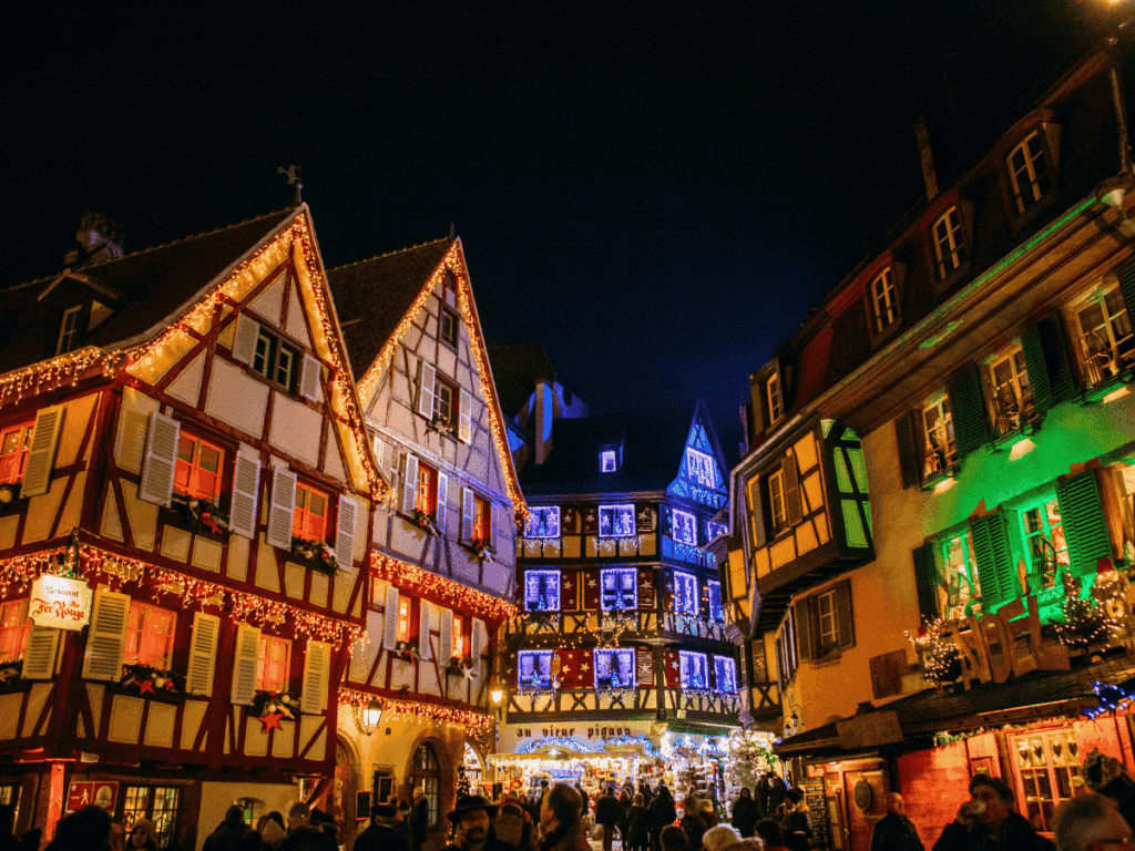 Half-timbered houses in Colmar at night, decorated with glowing Christmas lights or red, green, and blue. 