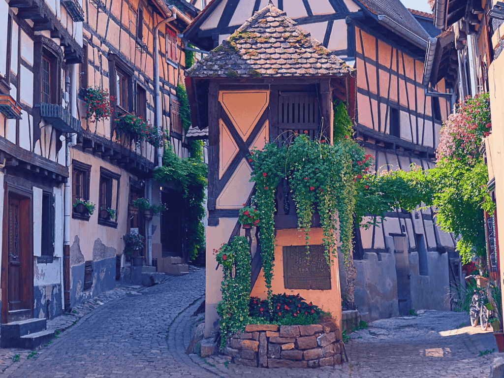 A narrow cobblestone alley in Riquewihr winds around charming half-timbered houses, with a small ivy-draped tower at the intersection blooming with red flowers.