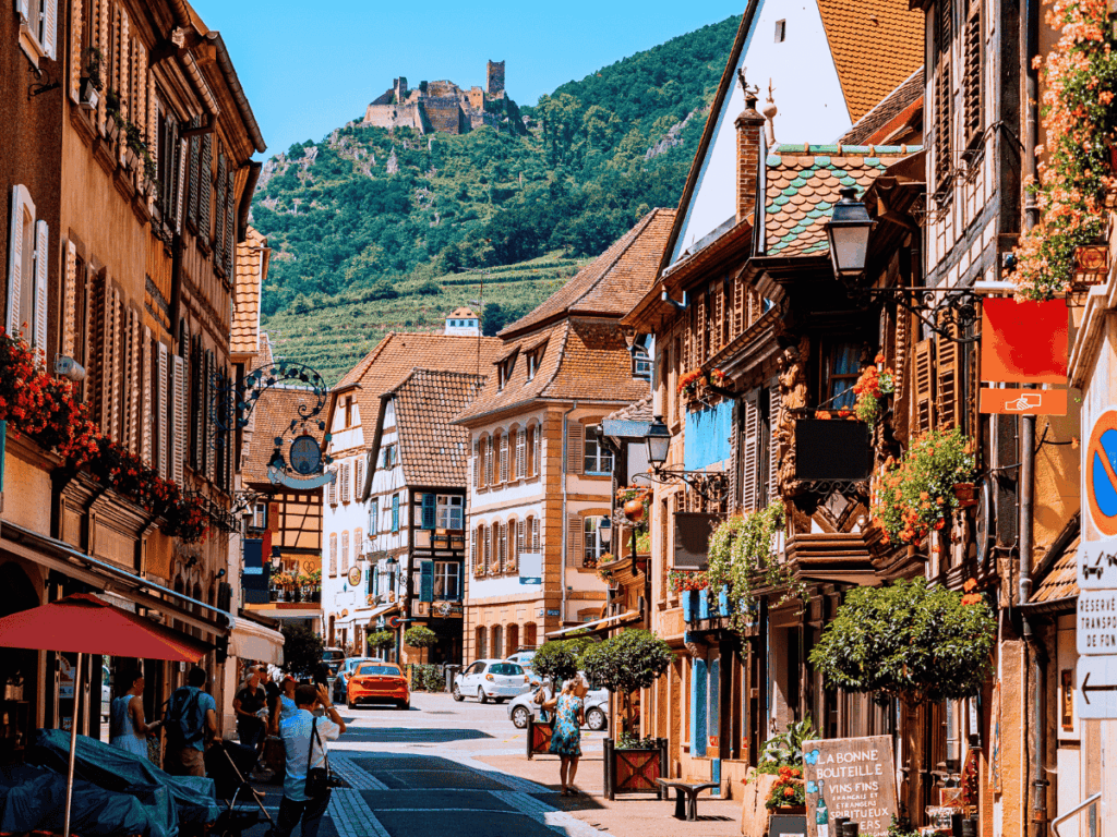 Colorful half-timbered buildings line a charming street in Ribeauvillé, France, with the ruins of Château de Saint-Ulrich perched on the lush green hill in the background.