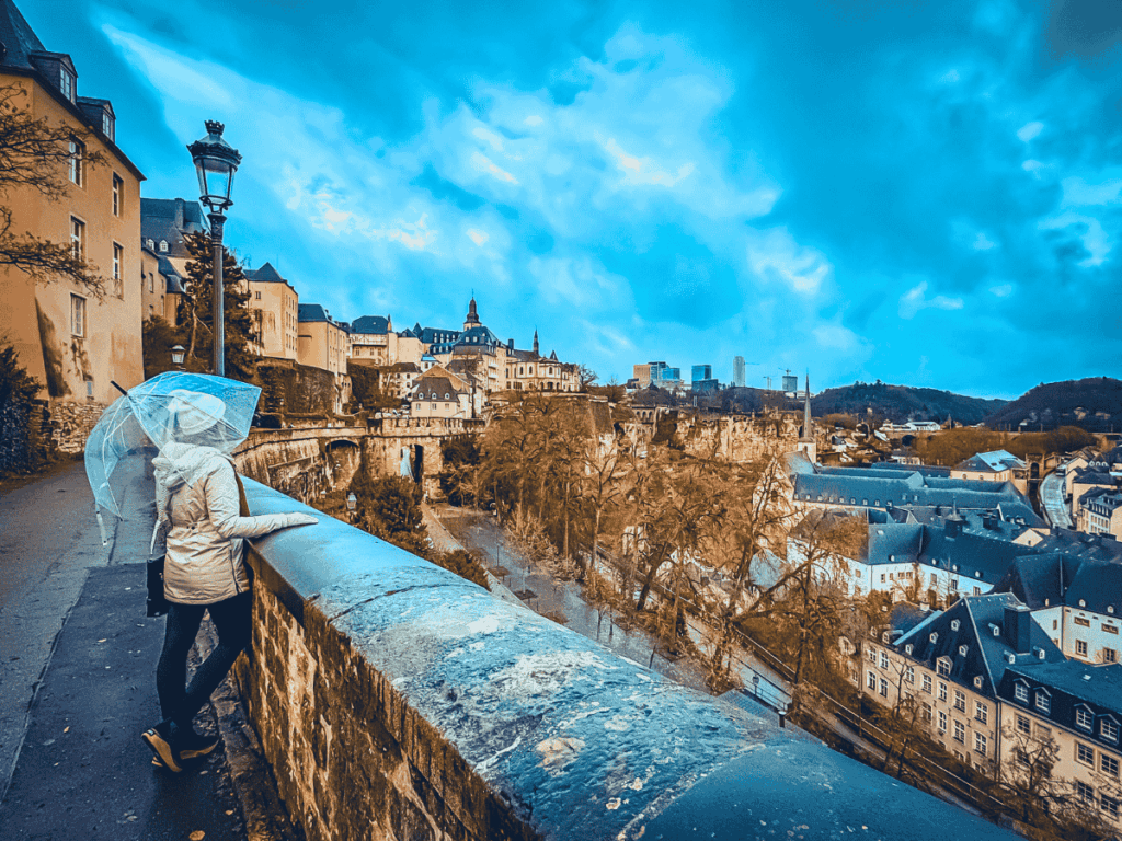 Kate holding a transparent umbrella looks out over the dramatic valley and layered cityscape of Luxembourg City from a stone terrace.