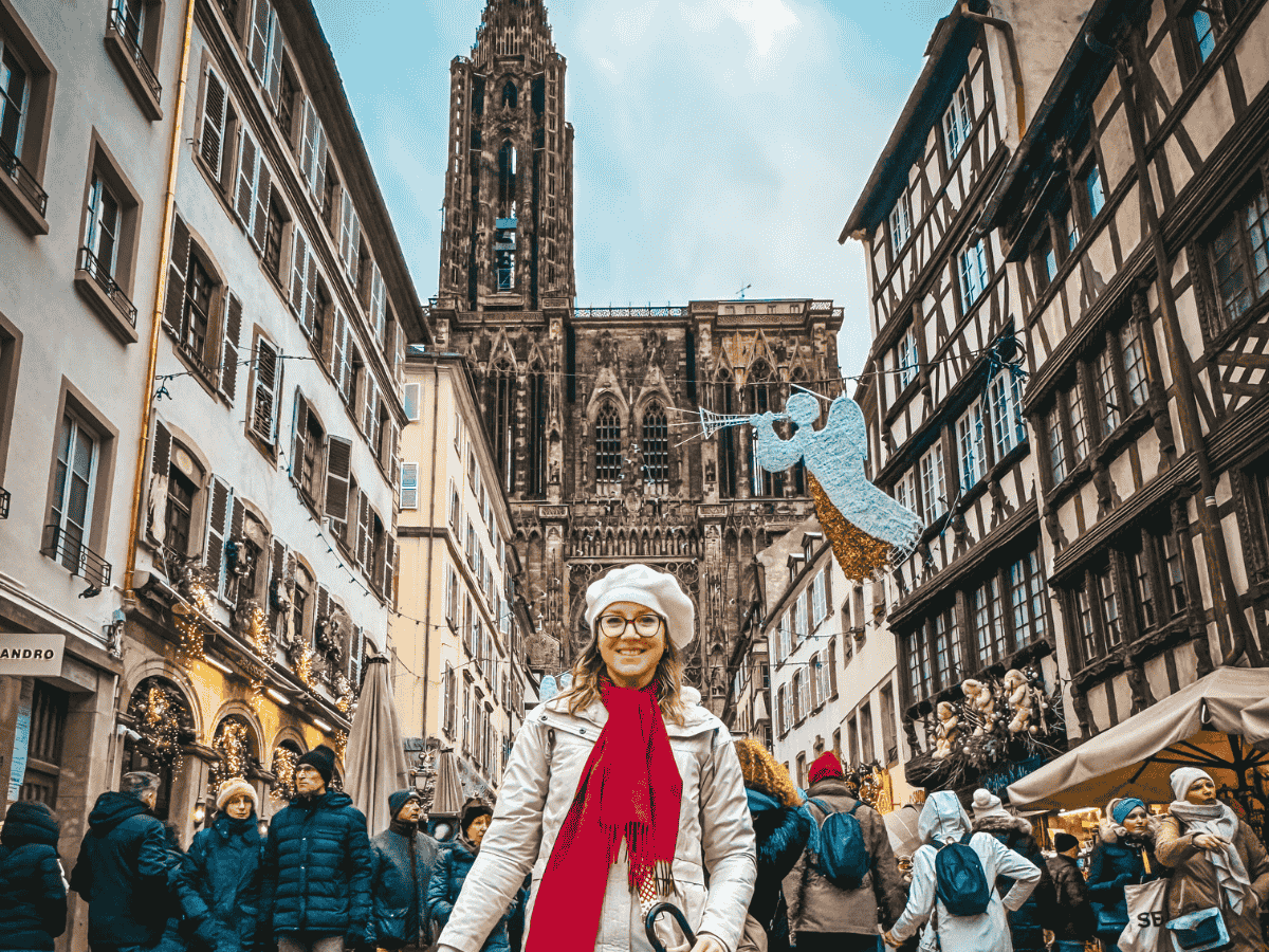 A smiling Kate, dressed in winter clothes, stands in front of the Strasbourg Cathedral, surrounded by festive holiday decorations and crowds enjoying the Christmas market atmosphere.