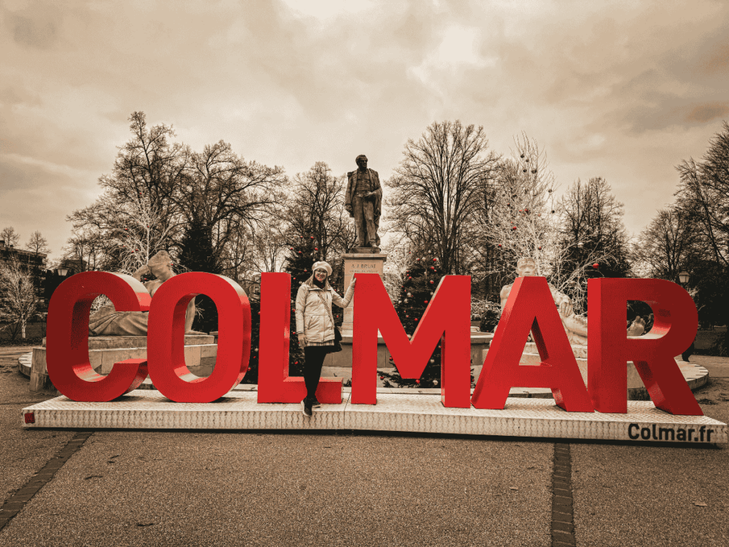 Kate stands inside the large red “COLMAR” sign installation in front of a statue and winter holiday decorations in a park in Colmar, France.