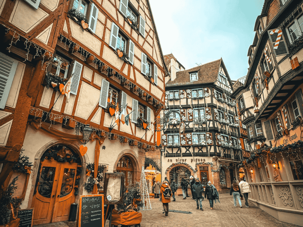 Charming street scene in Colmar, France, with people walking between ornately decorated half-timbered buildings.