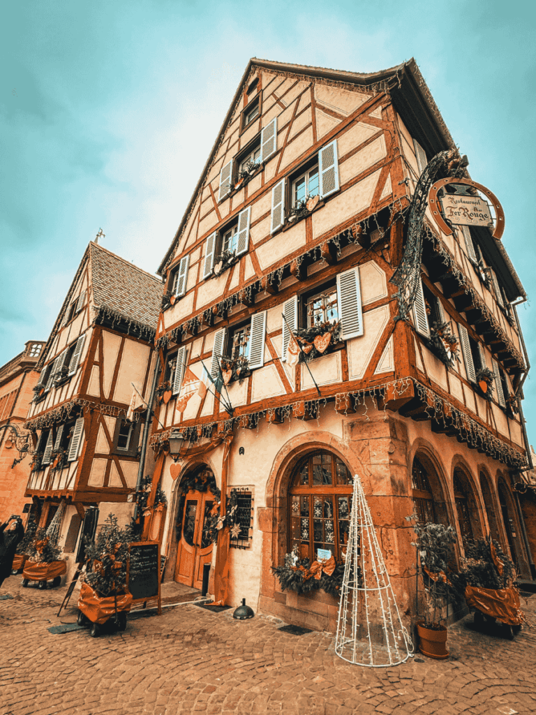 Festively adorned timber-framed building in Colmar housing the Restaurant Le Fer Rouge, with garlands, heart decorations, and a white wire Christmas tree outside.