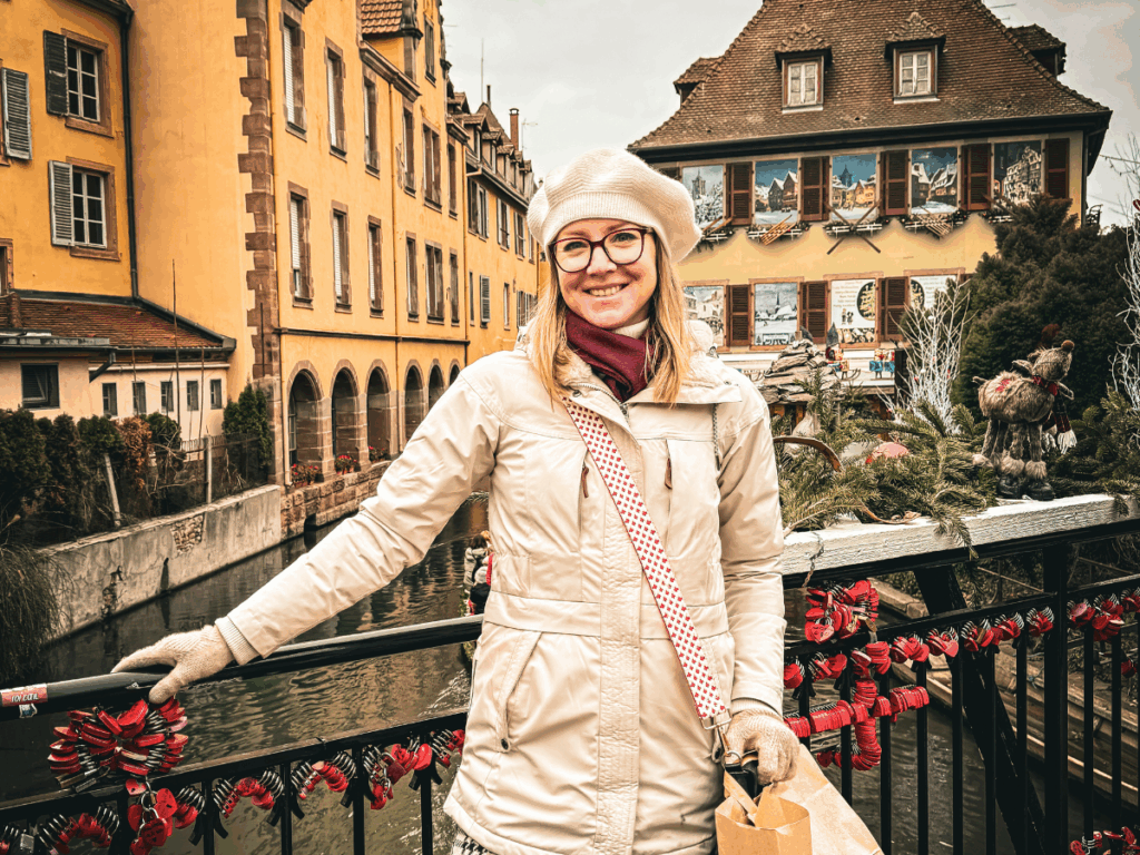 Kate smiles on a festive bridge in Colmar, France, surrounded by holiday decorations and colorful historic buildings along the canal.