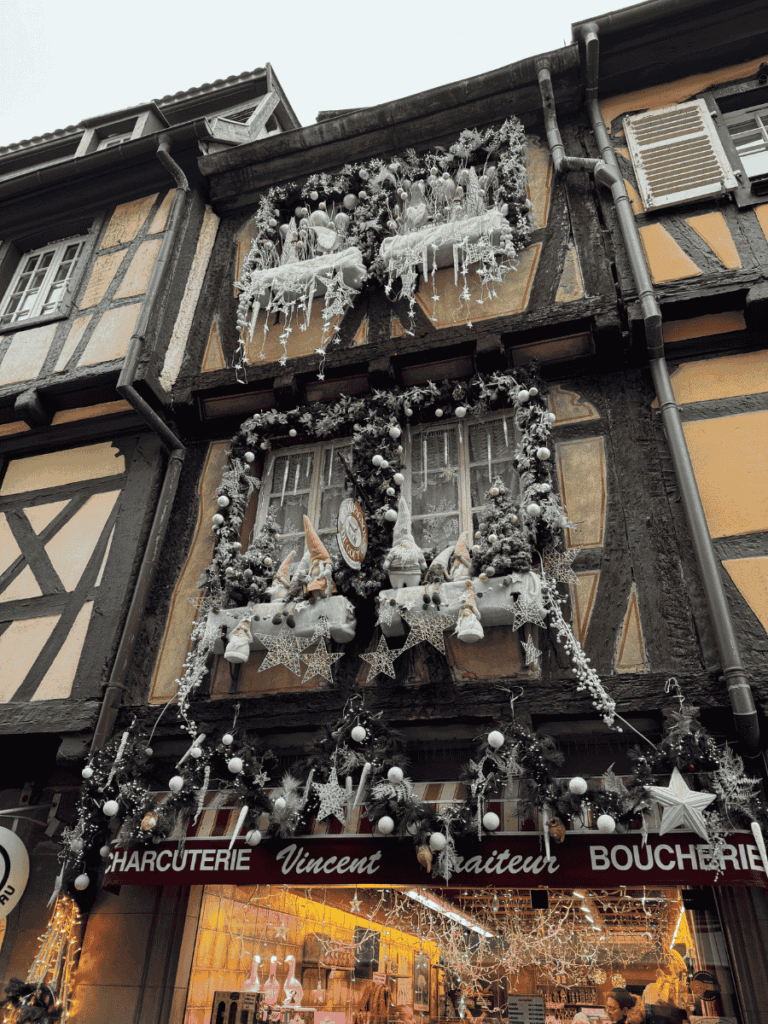 Festively decorated facade of Charcuterie Vincent in Colmar, with gnome figures, star ornaments, and garlands framing the windows of a half-timbered building.