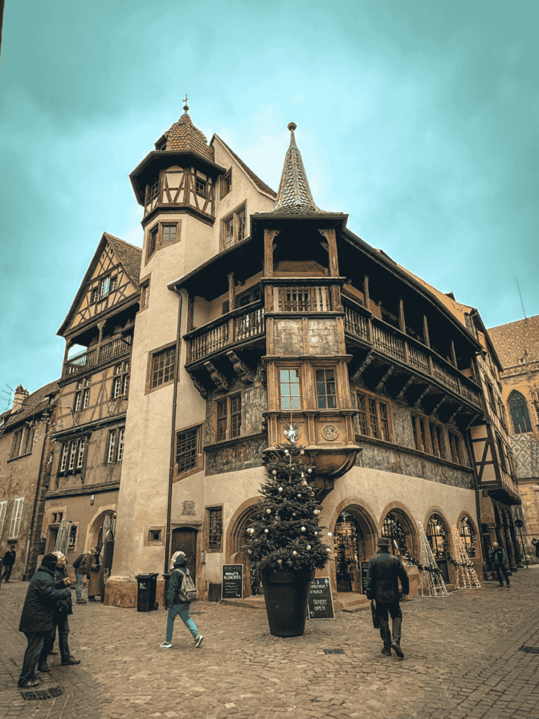 The Maison Pfister in Colmar, France, a striking Renaissance-era building with ornate wooden balconies and a decorated Christmas tree at its entrance.