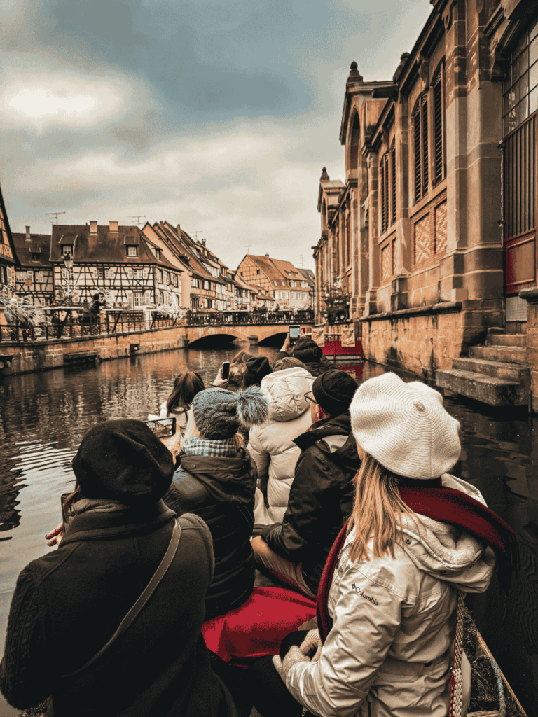 Kate sits at the edge of a boat with other bundled-up passengers during a canal tour in Colmar, passing by half-timbered houses and the historic covered market building.