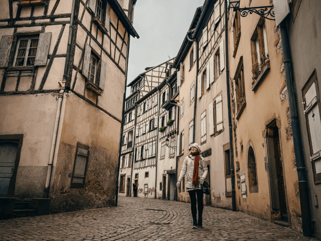 Kate walks down a quiet cobblestone street lined with pastel-colored half-timbered buildings in Colmar, France, looking up at the charming architecture.