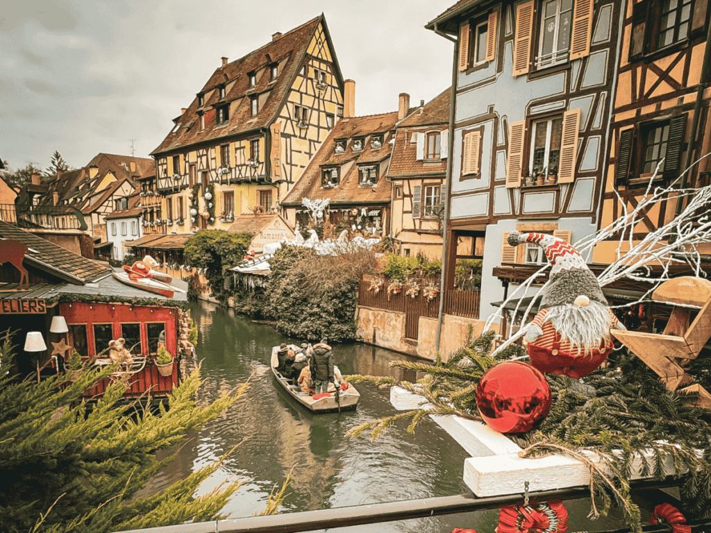 Festively decorated canal scene in Colmar’s Little Venice with a boat of tourists passing between colorful half-timbered buildings and Christmas ornaments in the foreground.