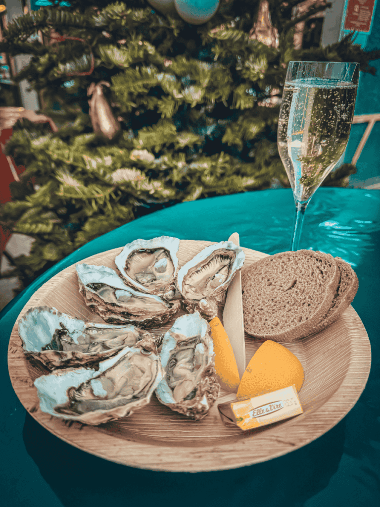 Close-up of a festive holiday table in Colmar featuring a wooden plate of fresh oysters with lemon wedges, rye bread, butter, and a glass of sparkling wine in front of a Christmas tree.