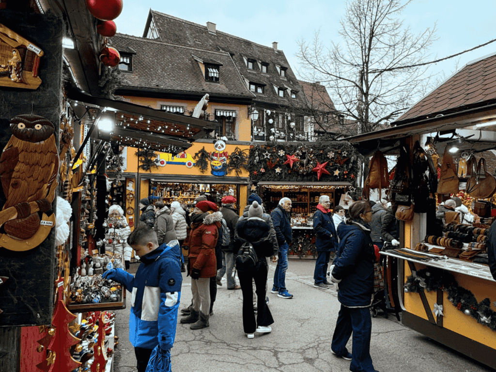 Crowds browse handcrafted ornaments, leather goods, and festive decor at the colorful outdoor stalls of the Colmar Christmas market.