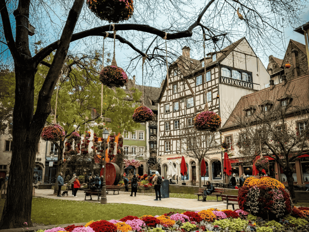 Colorful flowers and hanging floral displays brighten a charming square in Strasbourg, surrounded by half-timbered buildings and cozy cafés.