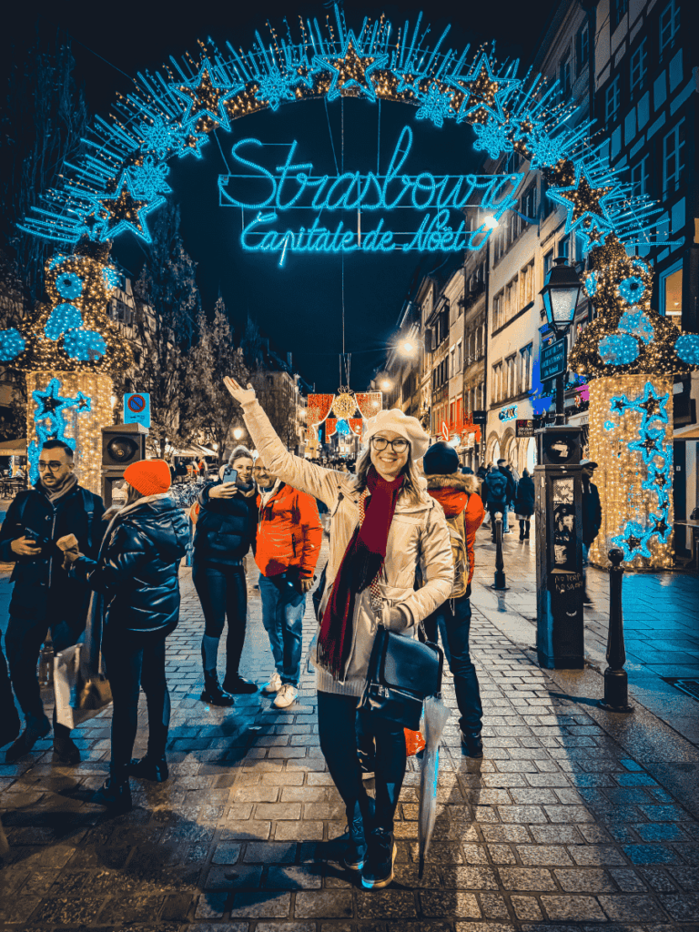 Kate stands beneath Strasbourg’s glowing “Capitale de Noël” Christmas light archway, smiling amid the festive evening crowds.