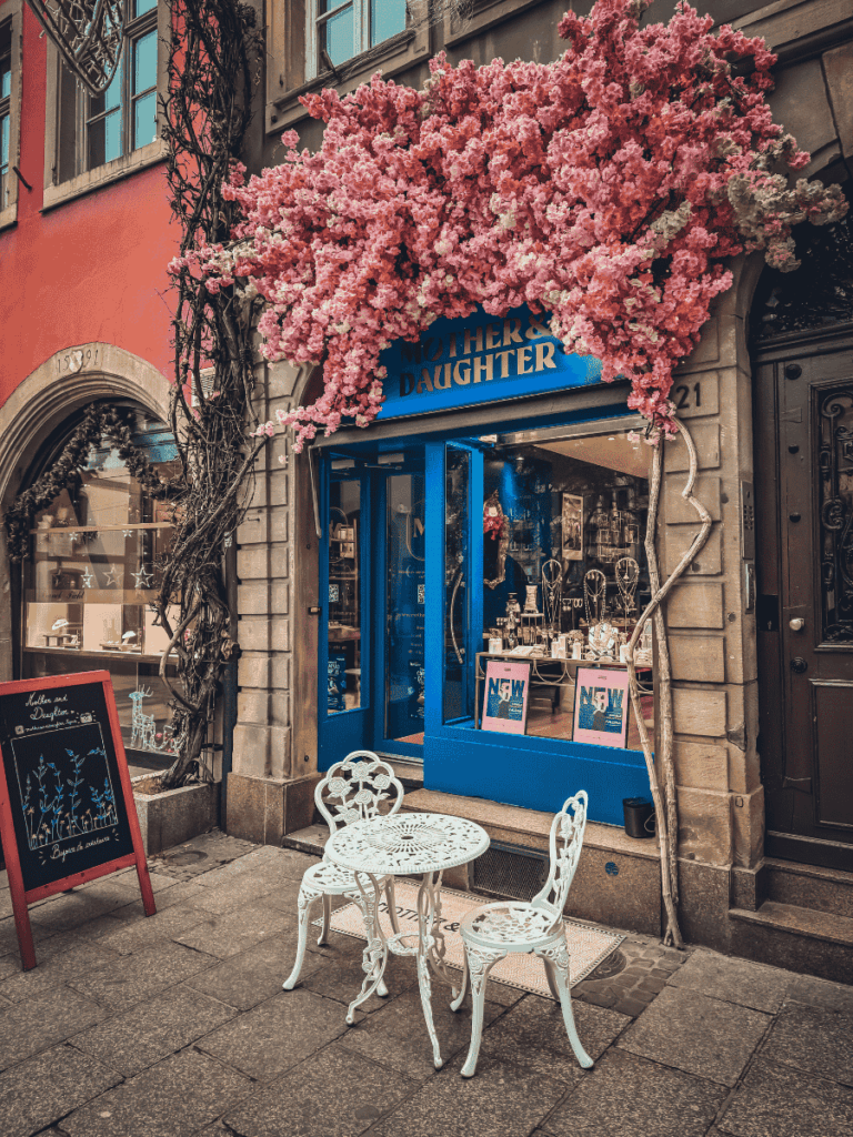 A charming Strasbourg boutique with a bright blue storefront is adorned with cascading pink flowers and a small white café table outside.
