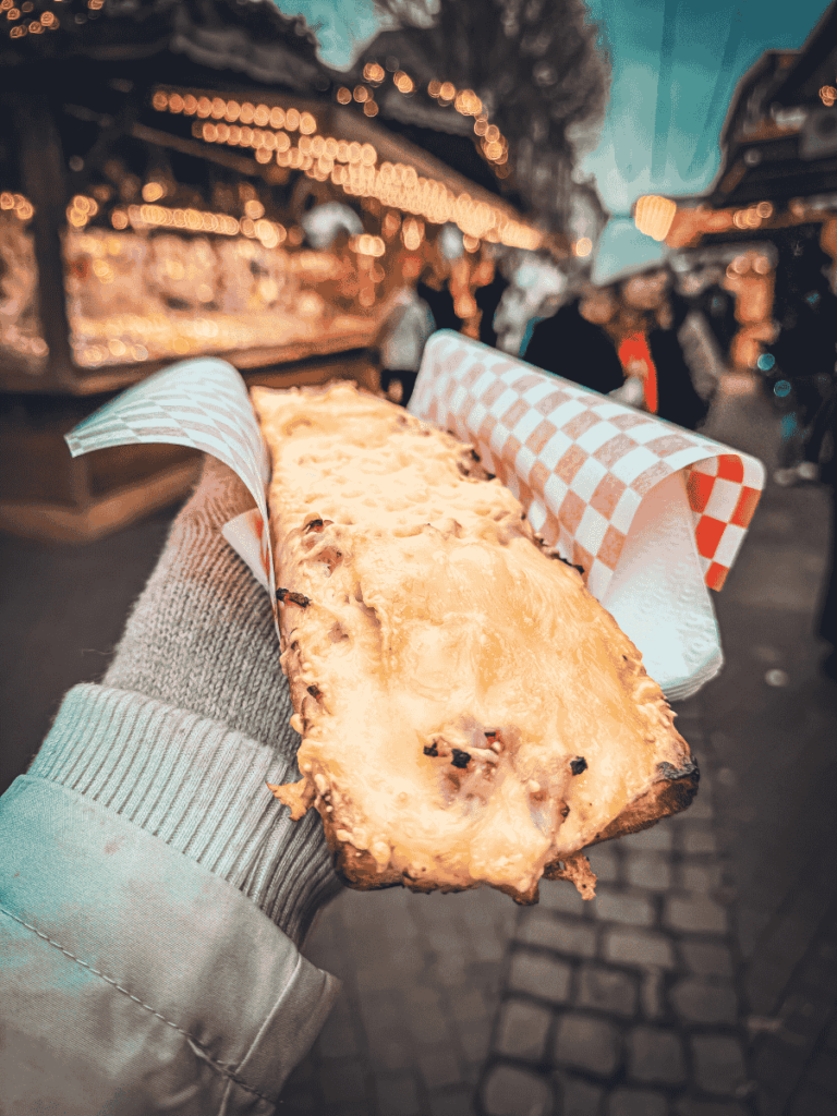 Strings of purple and white Christmas lights and ornaments illuminate a narrow street in Strasbourg filled with evening shoppers.

You said:
She Knows Alt Text said:

A hand holds a slice of melted cheese tartine at Strasbourg’s Christmas market with festive stalls glowing in the background.