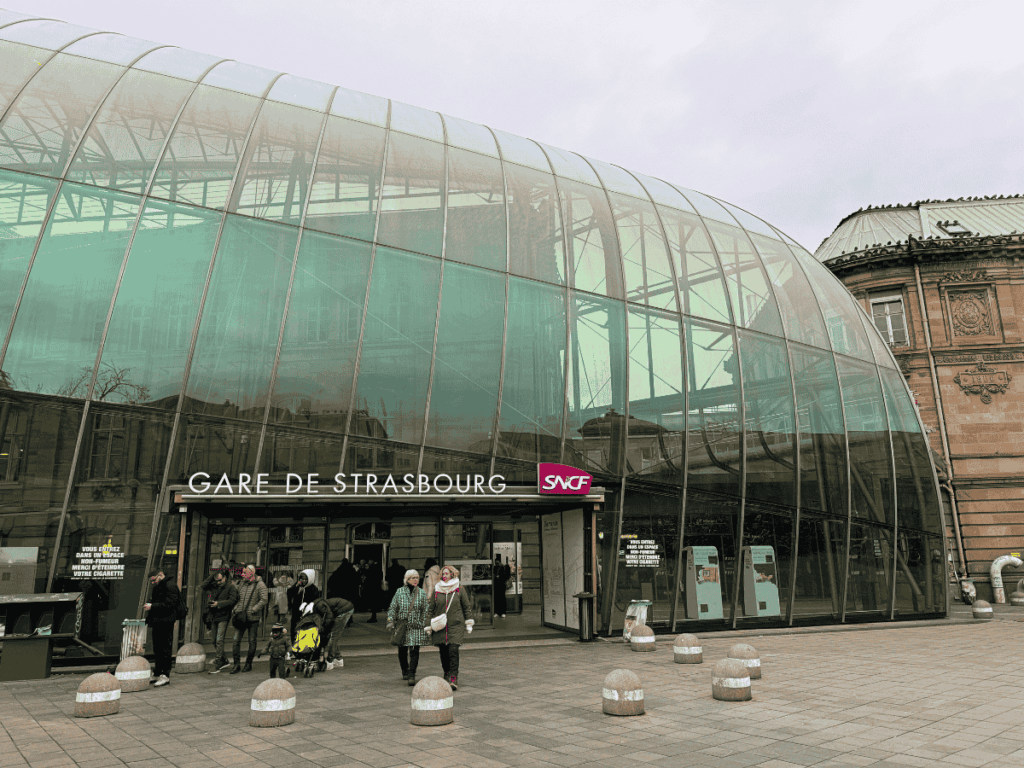 The glass-domed entrance of Gare de Strasbourg reflects the surrounding historic buildings as people walk in and out.