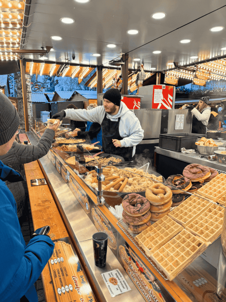 A vendor serves steaming food at a bustling Strasbourg Christmas market stall filled with pretzels, waffles, and festive lights.