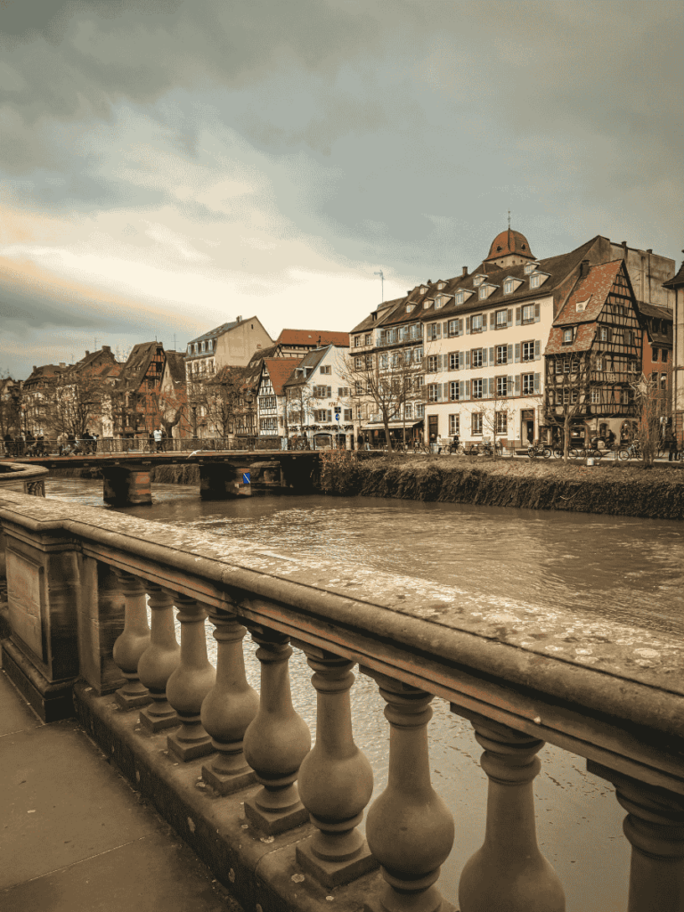 A view from a stone bridge in Strasbourg overlooks the Ill River and rows of traditional half-timbered houses under a cloudy sky.