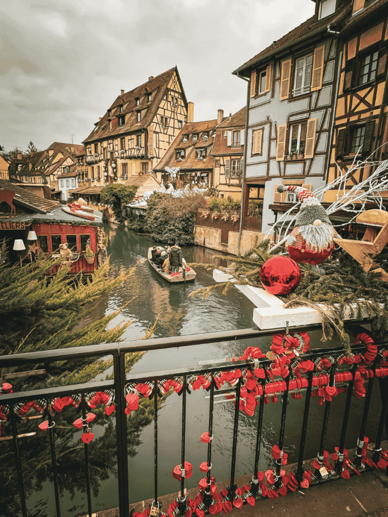 A festive canal scene in Colmar’s Little Venice shows a small boat gliding past half-timbered houses, with Christmas decorations and red heart locks adorning the bridge.
