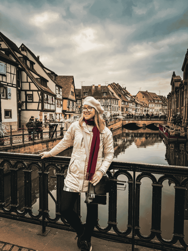 Kate, in a white coat and beret, smiles on a bridge in Colmar’s Little Venice, framed by the town’s iconic timbered buildings and a calm, reflective canal.