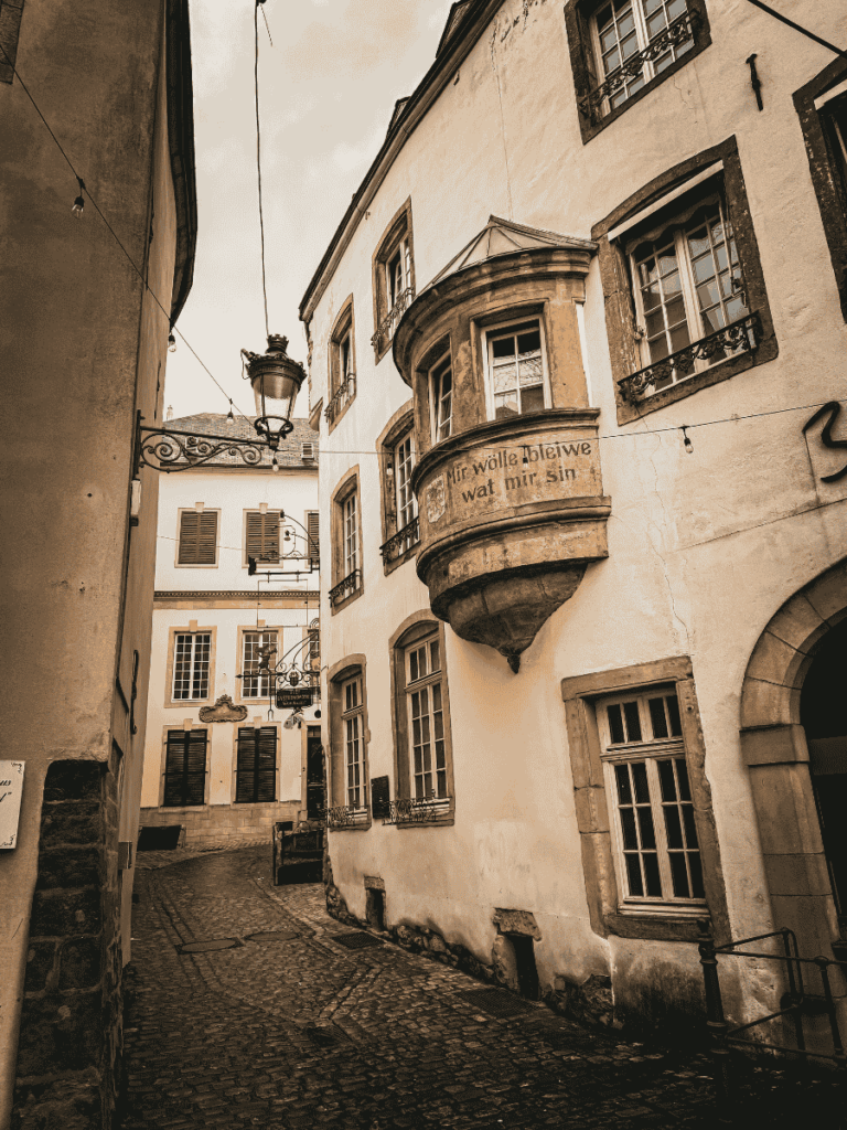 A narrow cobblestone alley in Luxembourg City winds past historic buildings, with a rounded bay window displaying the phrase “Mir wëlle bleiwe wat mir sin.”