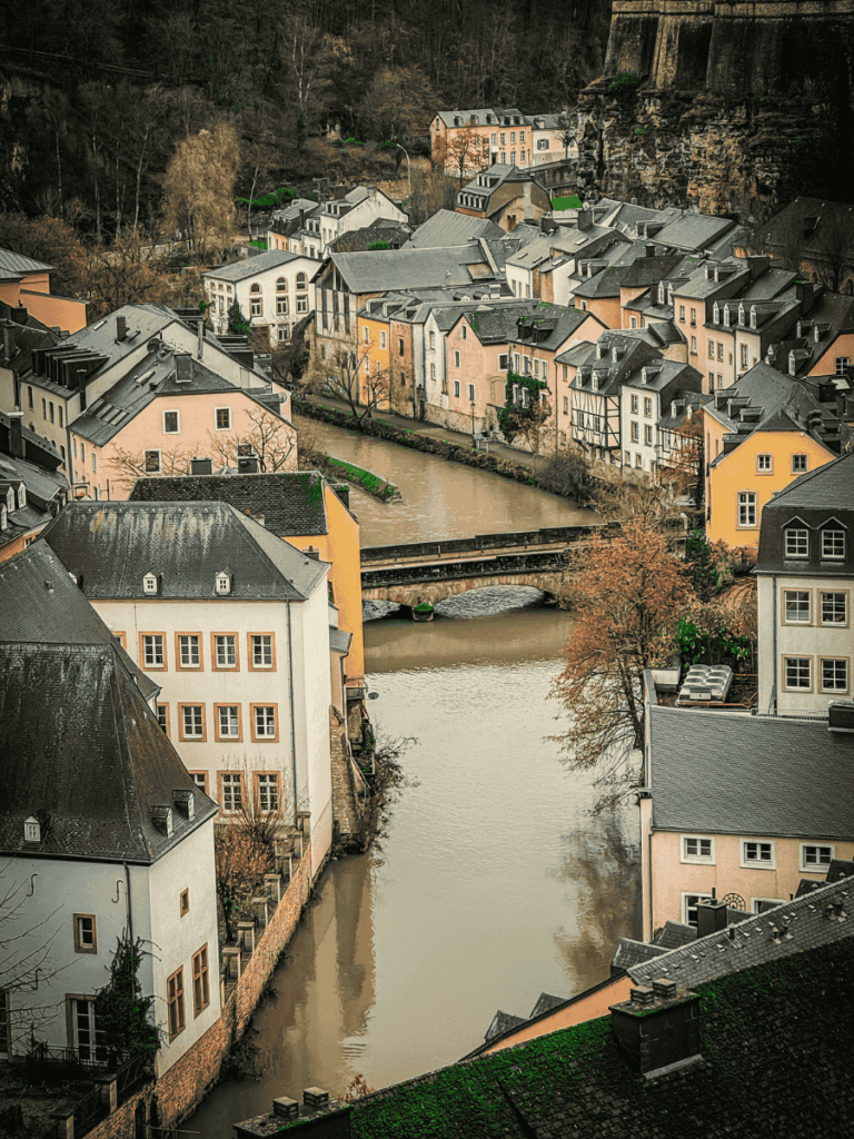 An aerial view of pastel-colored houses and winding streets along the Alzette River in the lower part of Luxembourg City, nestled against steep cliffs.