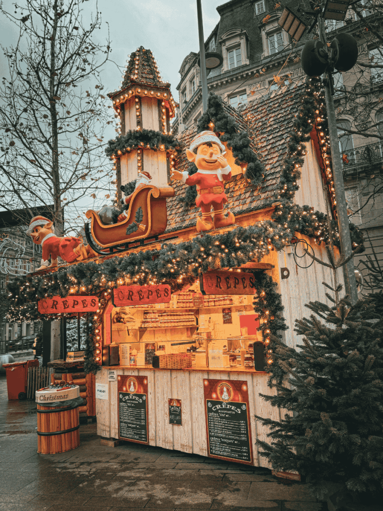 A festive wooden crêpe stall at the Luxembourg Christmas market is decorated with sparkling lights, garlands, cheerful elves, and Santa’s sleigh on the roof.