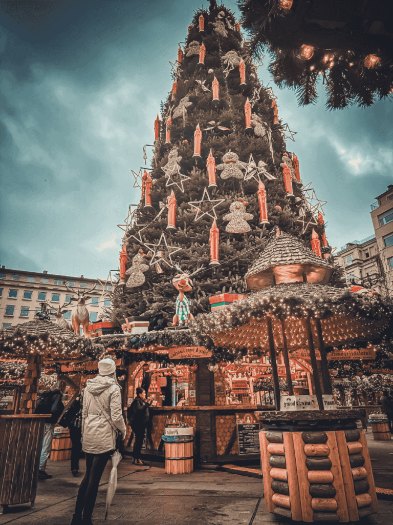 A towering Christmas tree adorned with glowing red candles, stars, and gingerbread figures stands at the heart of Luxembourg’s Christmas market, surrounded by cozy, illuminated wooden huts.