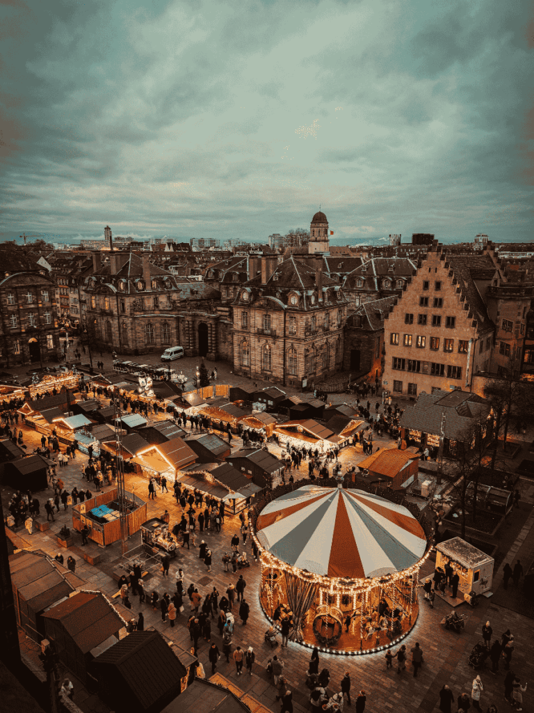 Aerial view of Strasbourg’s festive Christmas market with glowing wooden stalls and a carousel surrounded by historic buildings.