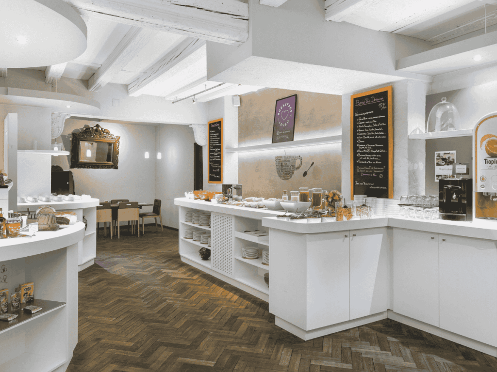 Bright breakfast buffet area at Hôtel de l'Europe by HappyCulture with white counters, fresh juices, cereals, pastries, and chalkboard menus on the wall.