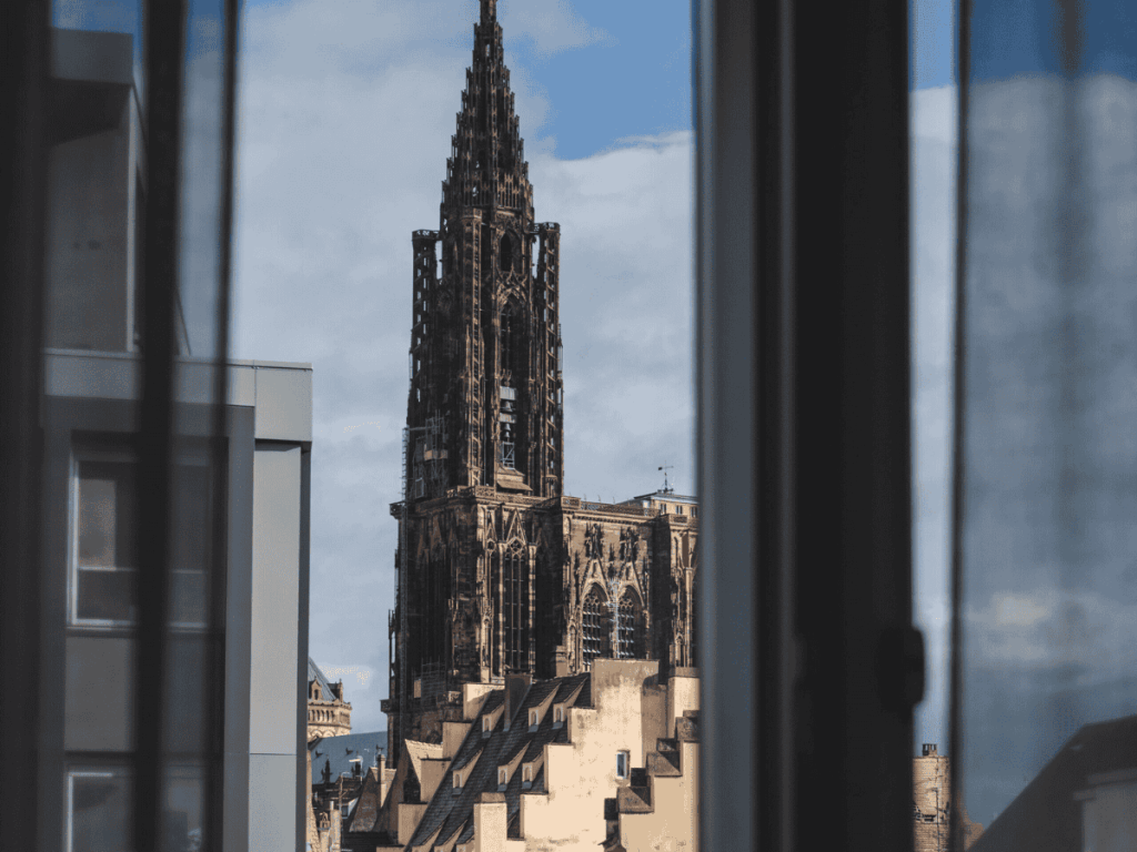 View of the Strasbourg Cathedral’s spire framed by modern and historic rooftops, seen through a hotel window at Maison Rouge Strasbourg Hotel & Spa.