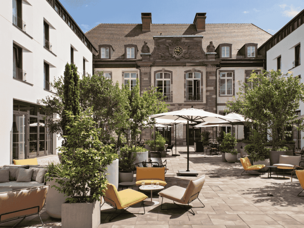 Sunlit courtyard at Hôtel LÉONOR featuring modern lounge seating, white parasols, and lush greenery set against the backdrop of a restored historic facade.