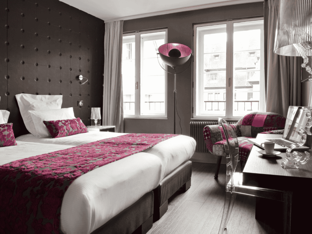 Chic hotel room at Hotel Rohan in Strasbourg featuring twin beds with fuchsia floral accents, a tufted dark wall, and eclectic decor including a patchwork armchair and transparent furniture.