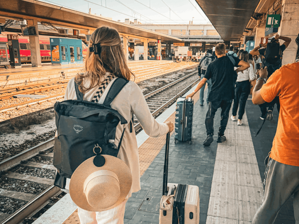 Kate walks along a busy European train platform with a suitcase in hand and a straw hat clipped to her backpack, surrounded by fellow travelers.