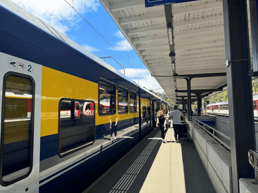 Travelers board a bright yellow and blue Bernese Oberland train under a sunny sky at a Swiss station platform.