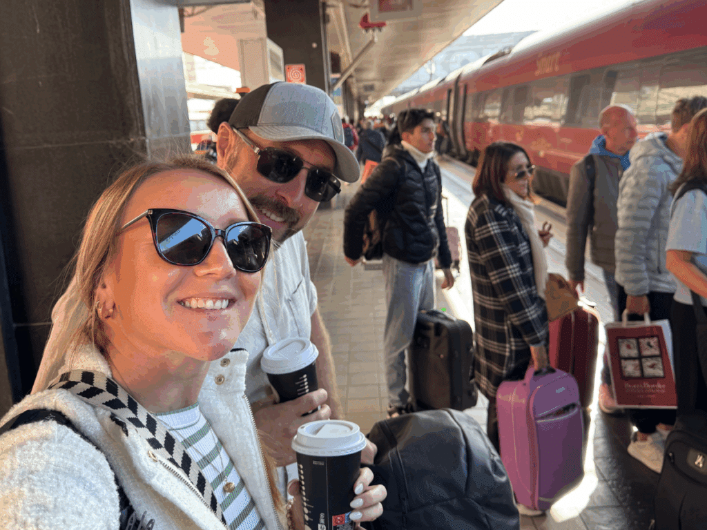 Kate and her travel companion smile with coffee cups in hand while waiting on a crowded platform beside a sleek red train.
