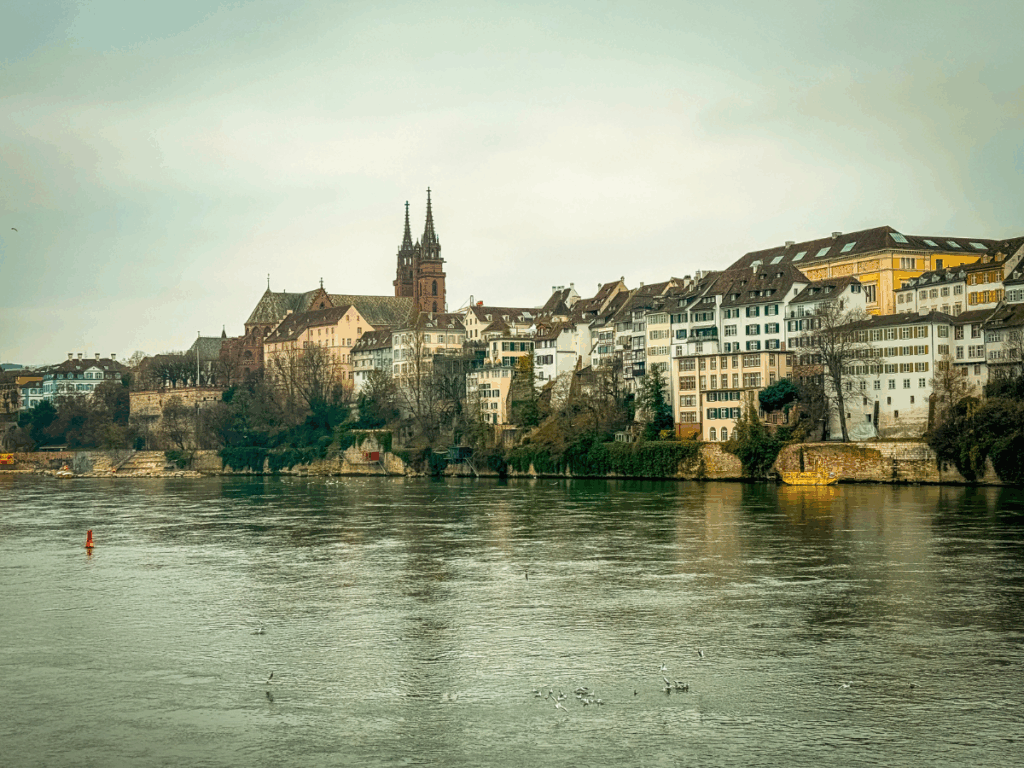 Colorful historic buildings and the red sandstone spires of Basel Minster line the Rhine River under a cloudy sky in Basel, Switzerland.