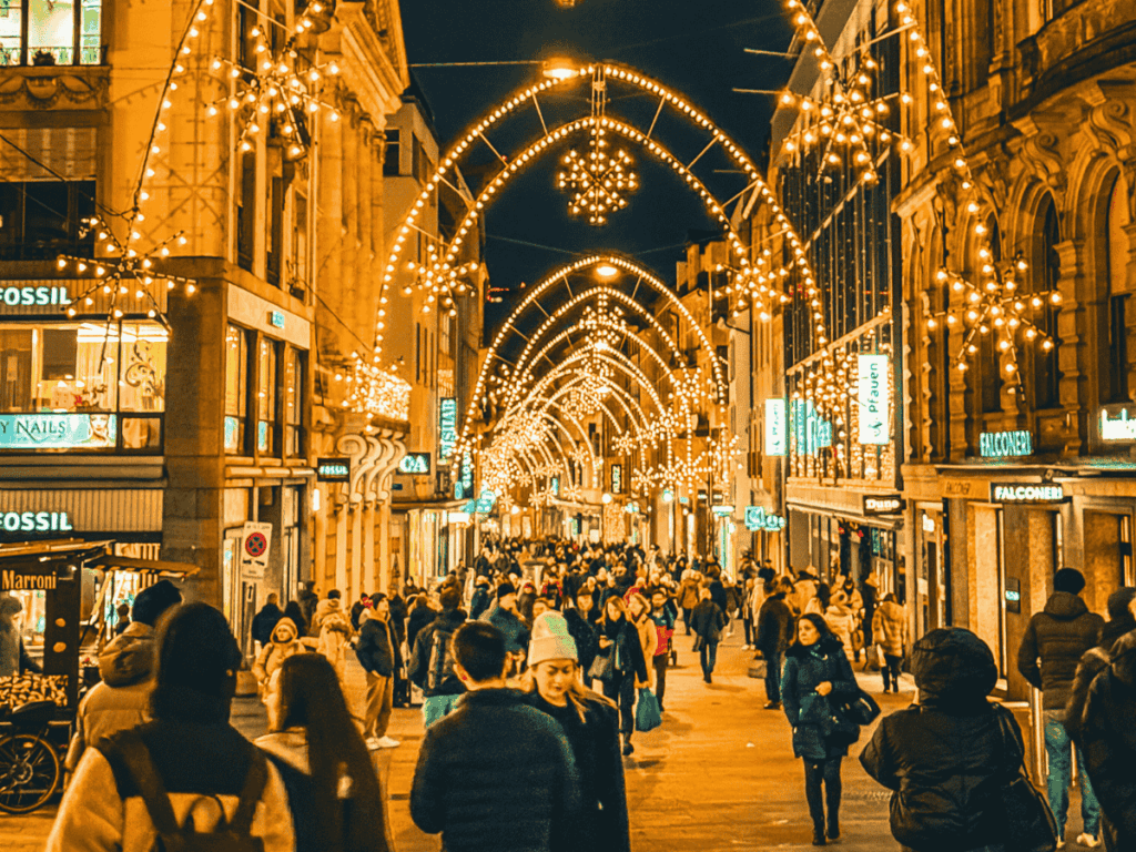 A bustling shopping street in Basel glows with golden holiday lights arching overhead, as crowds in winter coats stroll beneath the festive decorations.