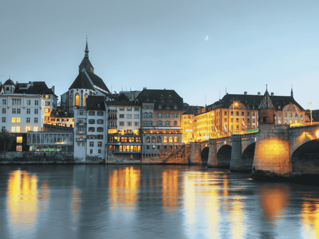 Evening lights from historic buildings and the stone Mittlere Brücke reflect off the calm Rhine River in Basel, with a crescent moon rising in the sky.