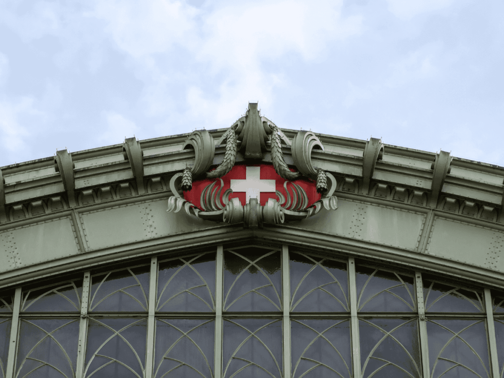 An ornate green metal facade features the Swiss flag emblem at the peak, set above arched windows with decorative glasswork.