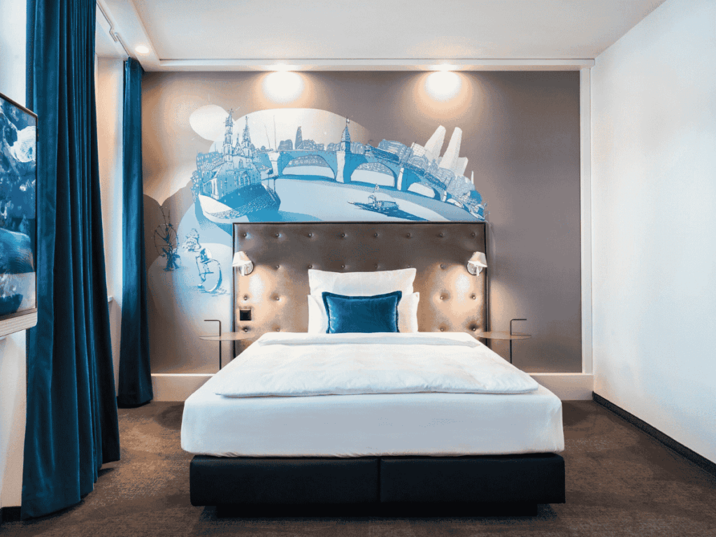 A contemporary hotel room in Basel is decorated with a cityscape mural above the bed, accented by deep blue curtains and a matching pillow.