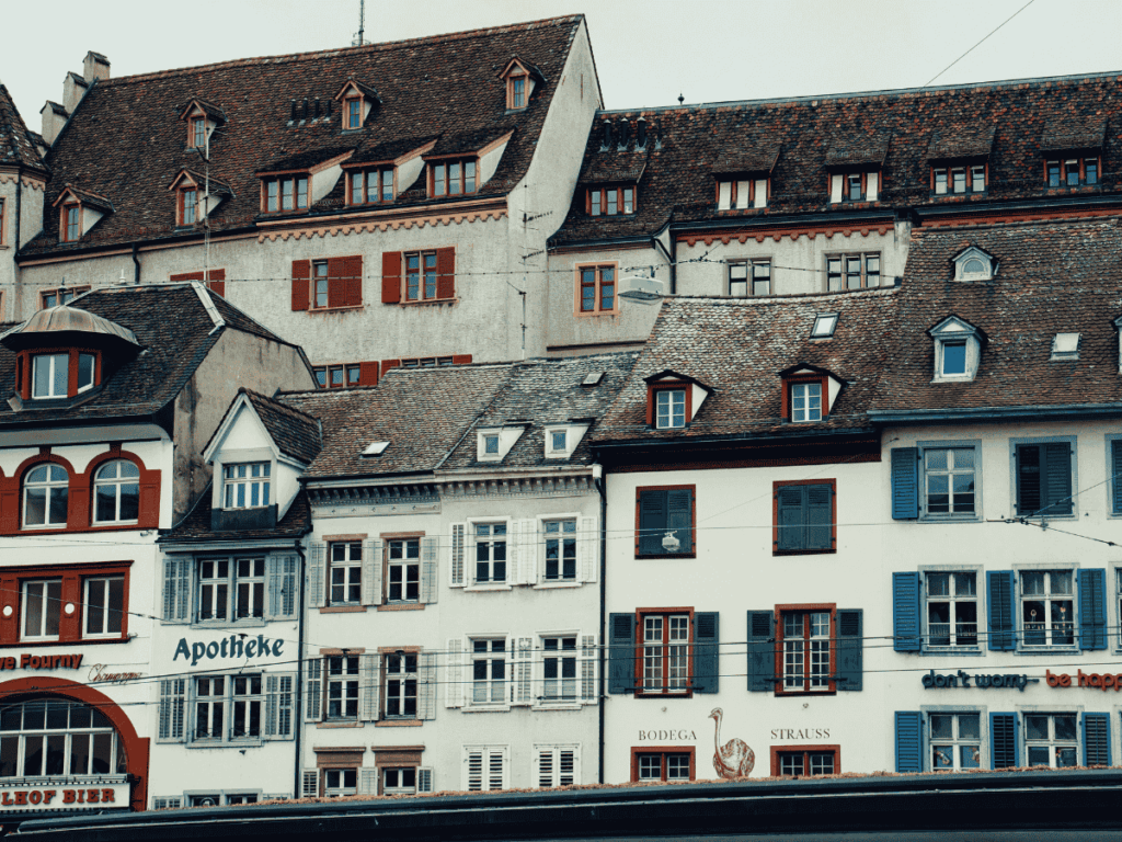 A row of colorful, tightly packed historic buildings in Basel features shops like a pharmacy and cafés, with classic shuttered windows and steep tiled roofs.