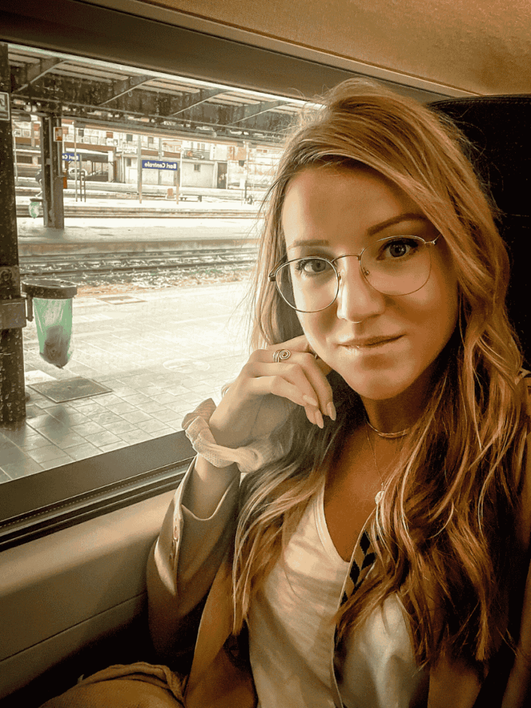 Kate sits by a train window with a soft smile, wearing glasses and a neutral-toned outfit, as the platform outside the station in Bari Centrale is visible in the background.
