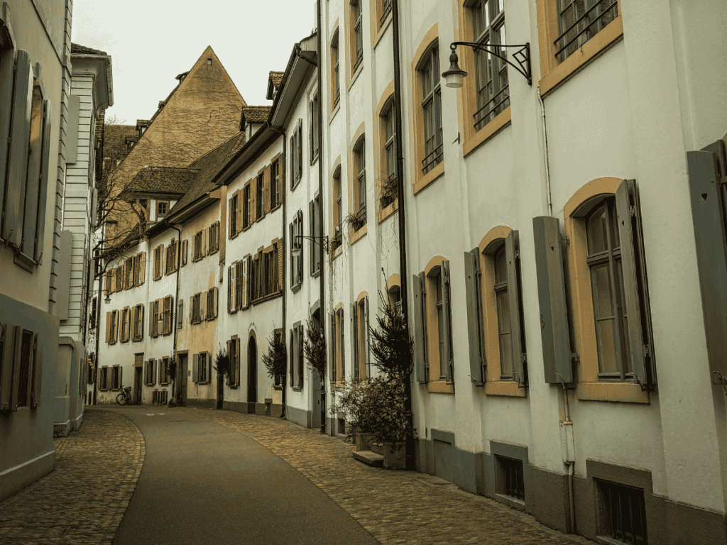A narrow, winding street in Basel is flanked by pastel-colored buildings with wooden shutters and potted plants lining the cobblestone path.