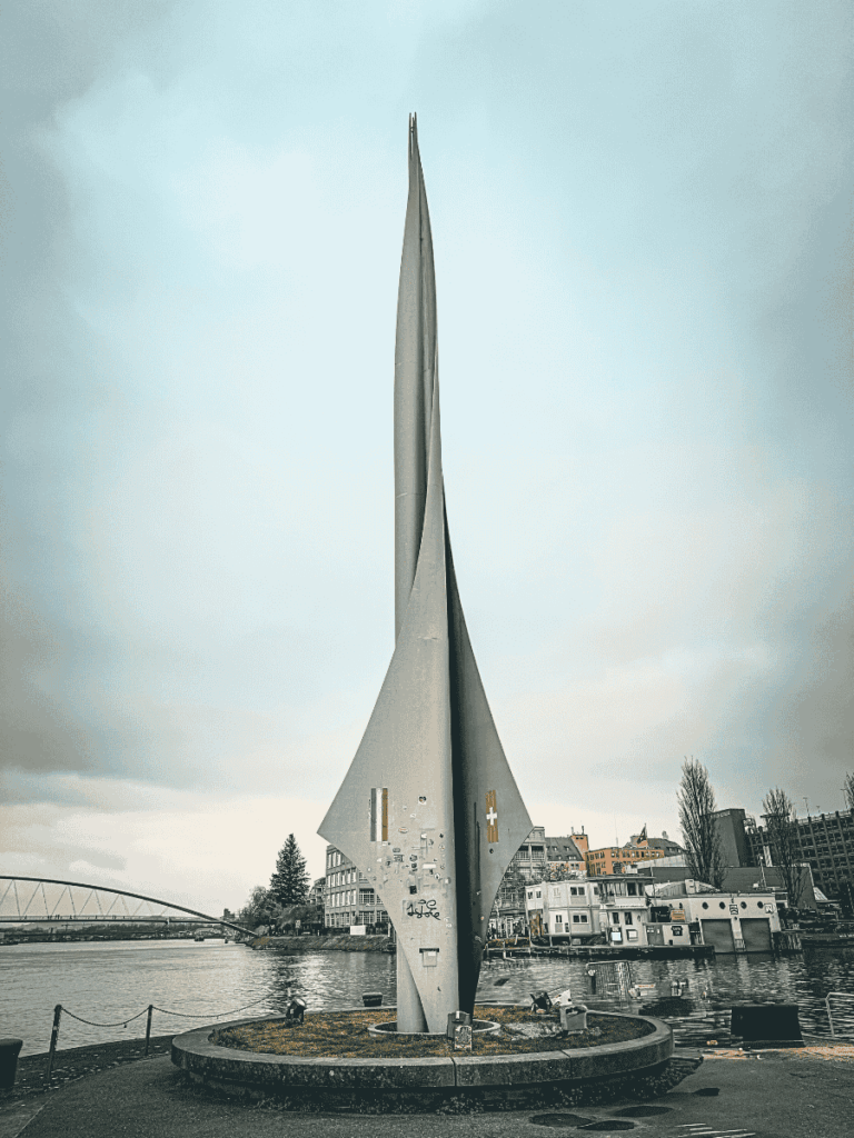 A tall, abstract steel sculpture with a sleek, curved form stands on the banks of the Rhine River in Basel, Switzerland.