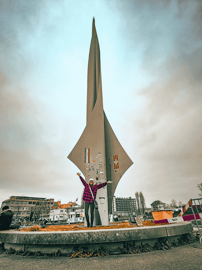 Kate stands joyfully with arms raised in front of a towering modern sculpture by the Rhine River in Basel, wearing a purple plaid jacket and winter hat.