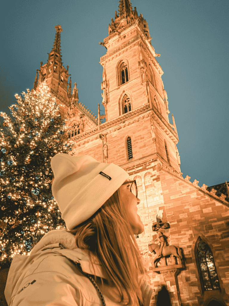 At night, Kate gazes up at the illuminated Basel Minster beside a glowing Christmas tree, bundled in a white hat and coat.