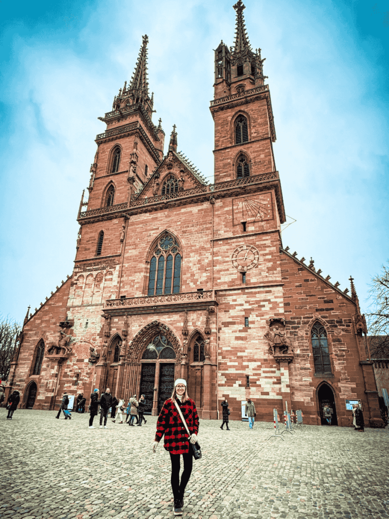 Kate stands smiling in front of the grand red sandstone Basel Minster, with its twin Gothic towers rising above the cobblestone square.